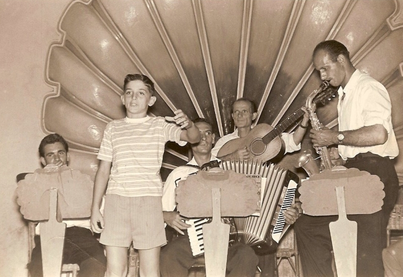 Il giovanissimo Antonio Volpe canta "Nel blu dipinto, di blu" accompagnato dall'orchestra sul palco a conchiglia del Dopolavoro durante una festa musicale